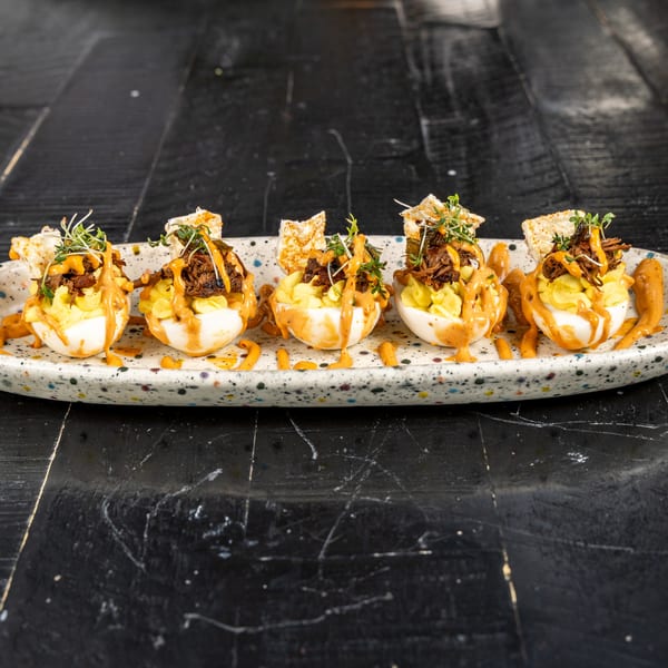 Five deviled eggs with pulled pork, sauce, microgreens, and tortilla chips on a speckled plate.
