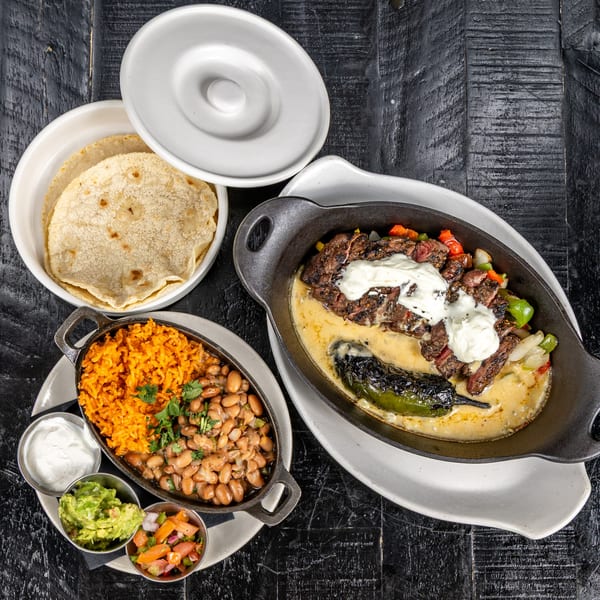 Mexican fajitas with steak, peppers, onions, cheese sauce, rice, beans, guacamole, and tortillas.