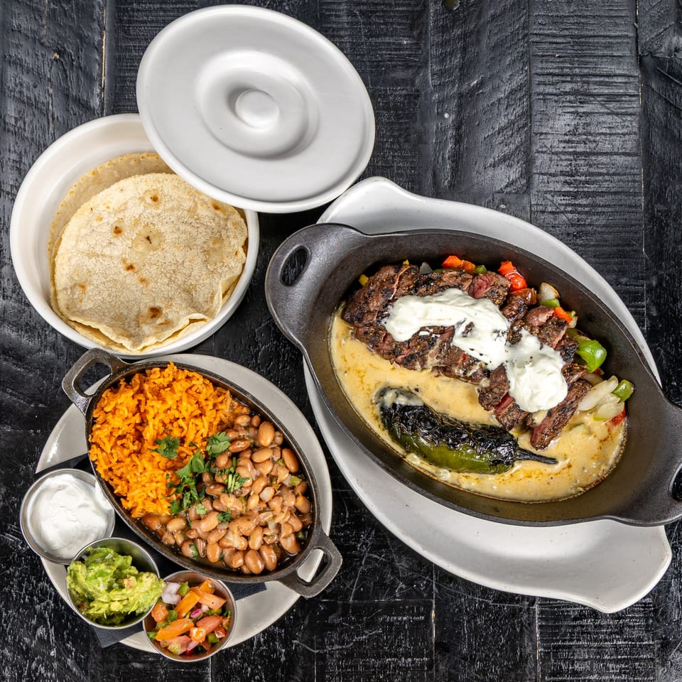 Mexican fajitas with steak, peppers, onions, cheese sauce, rice, beans, guacamole, and tortillas.