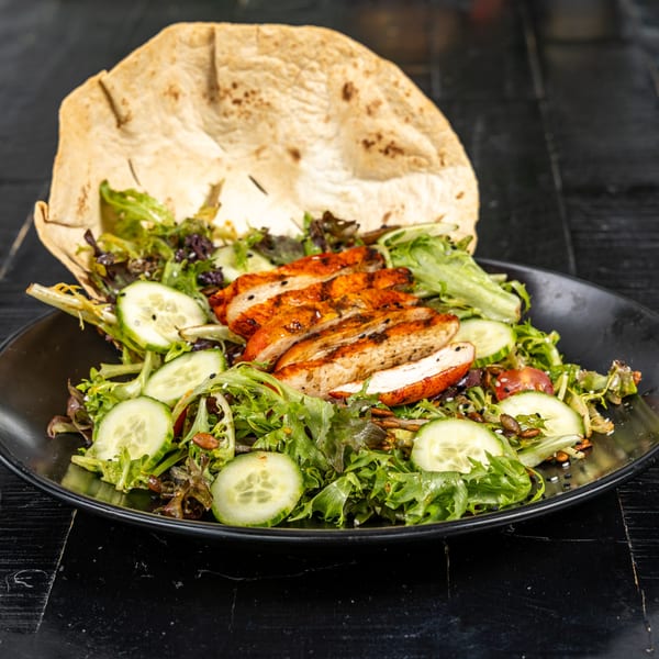 Grilled chicken salad with mixed greens, cucumbers, seeds, and a large tortilla on a black plate.