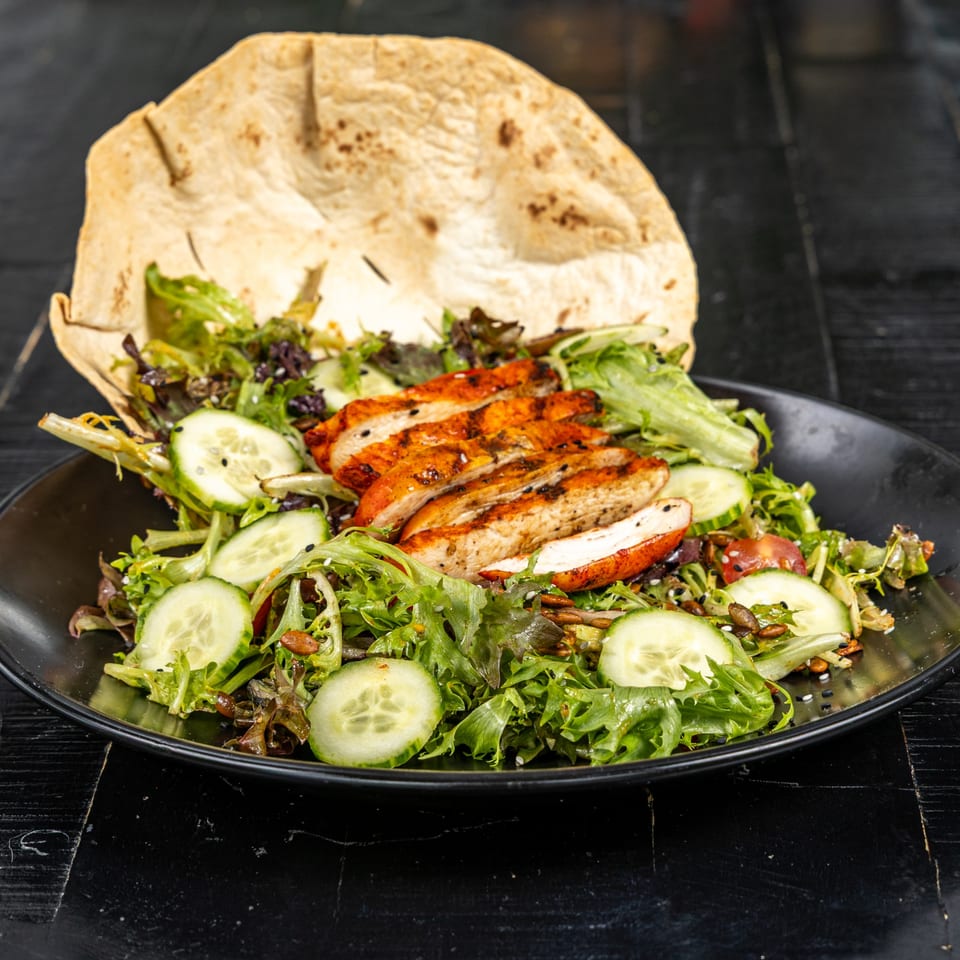 Grilled chicken salad with mixed greens, cucumbers, seeds, and a large tortilla on a black plate.