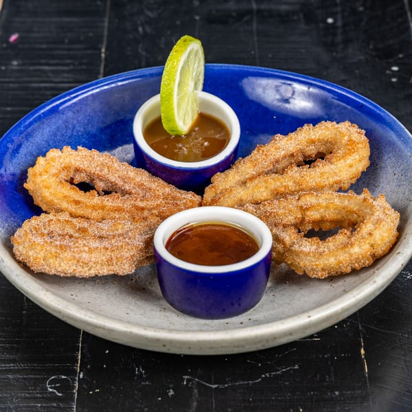 Four churros dusted with cinnamon sugar, served with two small bowls of caramel sauce and a lime wedge.