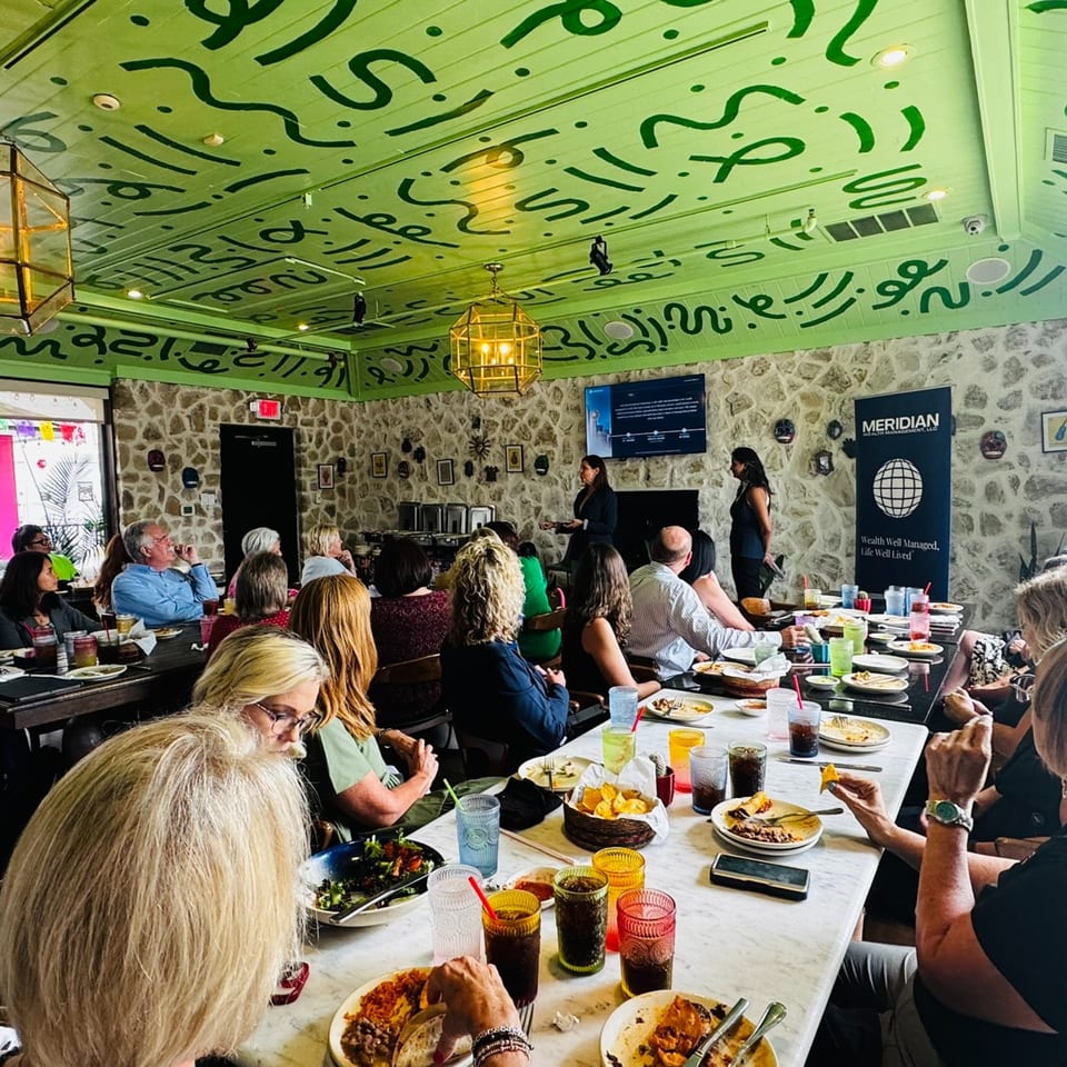 People seated at tables with food and drinks, listening to a presentation in a vibrant Mexican restaurant.