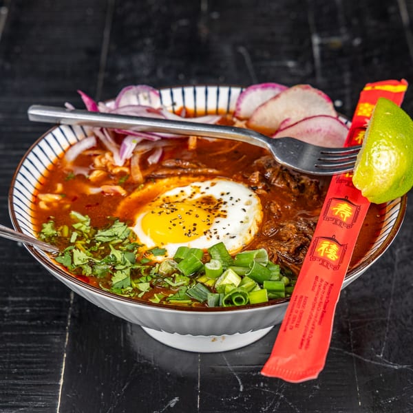 A bowl of Mexican birria with a fried egg, cilantro, green onions, radishes, and lime.