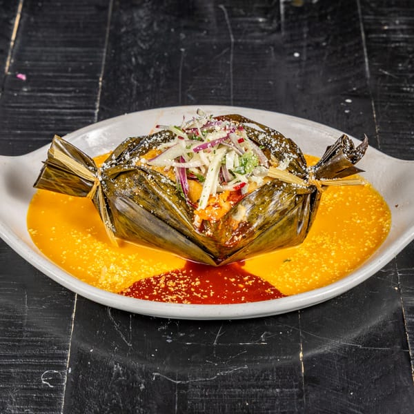 A Mexican tamale, wrapped in a corn husk, topped with slaw, on a plate with yellow and red sauces.