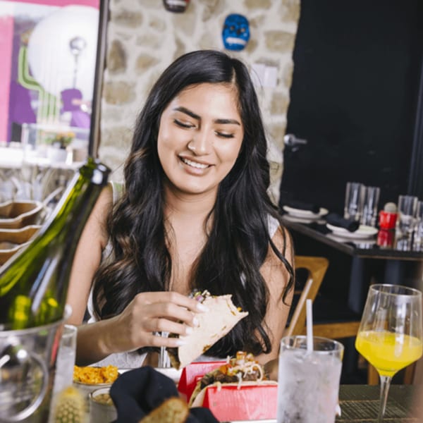 A smiling woman enjoying a taco at Neon Cactus, with drinks and a wine bottle on the table.